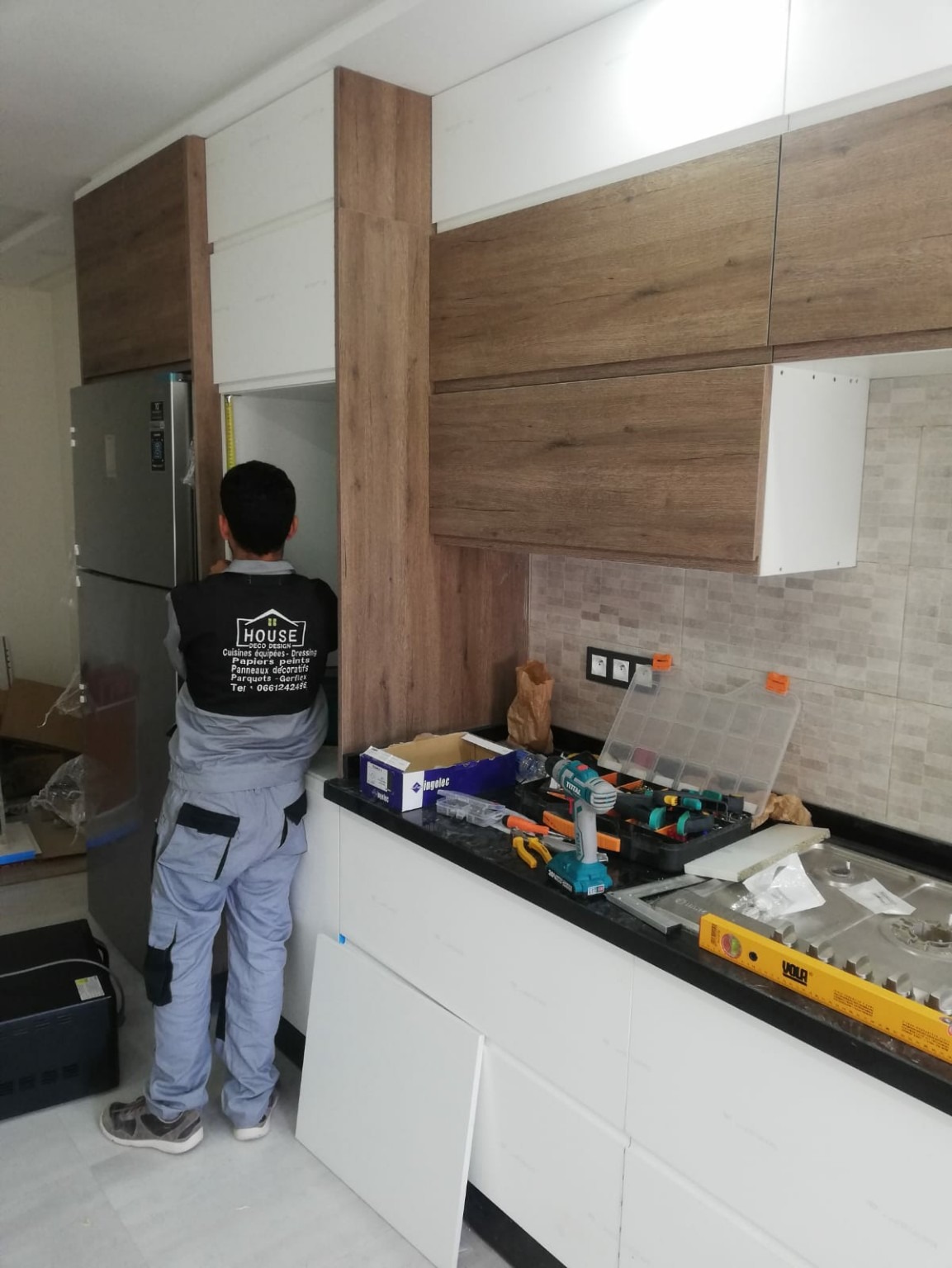 Pose de meubles de cuisine par une personne portant un vêtement House ; chantier en cours.
